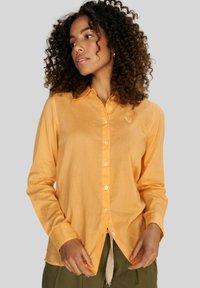 Camisa de manga larga en tejido color naranja claro con cuello, botones blancos y un sutil logo de corazón en el pecho. Ajuste holgado.