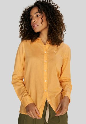 Camisa de manga larga en tejido color naranja claro con cuello, botones blancos y un sutil logo de corazón en el pecho. Ajuste holgado.