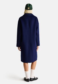 Cappotto di lana blu navy con vestibilità rilassata, maniche lunghe e colletto. Indossato con un berretto verde e scarpe nere, abbinato a calzini bianchi a coste.