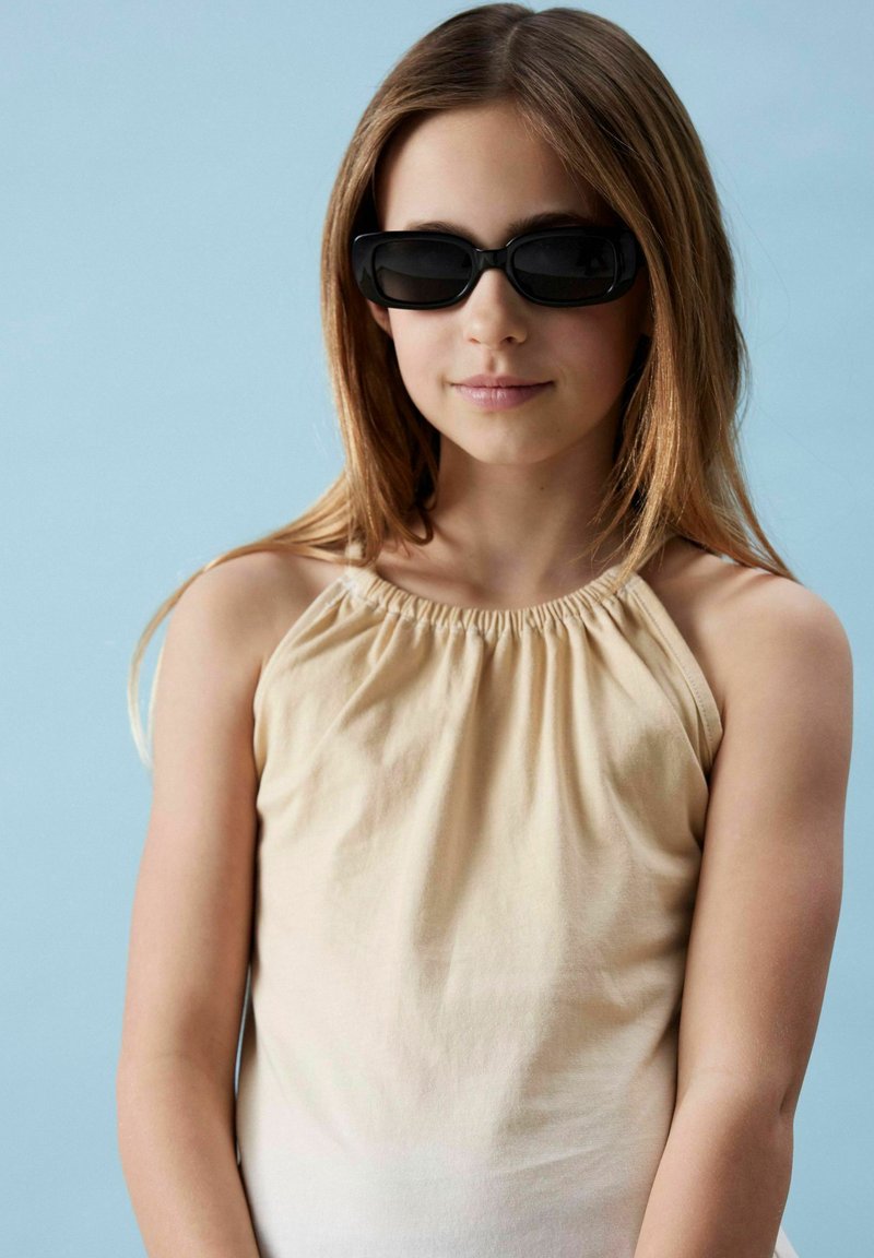 Top sans manches beige clair avec un col froncé, associé à des lunettes de soleil noires rectangulaires. Tissu lisse sur un fond bleu pâle.
