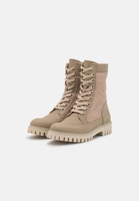 Beige Stiefeletten mit einer Mischung aus glattem und strukturiertem Stoff, ausgestattet mit einer Schnürung an der Vorderseite und einer kräftigen Gummisohle mit ausgeprägten Rillen.