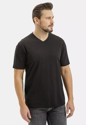 Mężczyzna o krótkich ciemnych włosach i brodzie, ubrany w czarną koszulkę z dekoltem w serek i szare dżinsy, stoi i patrzy w prawo na jednolitym tle.