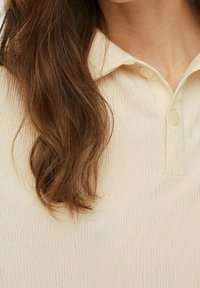 Camisa de color beige claro con cuello, confeccionada en una tela texturizada, que presenta un cierre de botón. La superficie arrugada añade dimensión a la prenda.