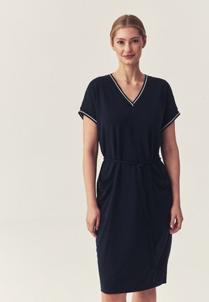 Frau trägt ein schwarzes knielanges Kleid mit kurzen Ärmeln, weißem Besatz am Ausschnitt und an den Ärmeln sowie einem gebundenen Taillengürtel.