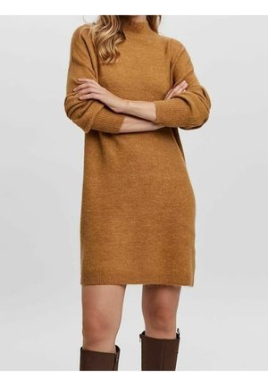 Kvinde iført en brun langærmet sweaterkjole og brune knæhøje støvler med armene korsede mod en ensartet lys baggrund.