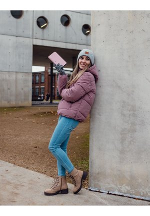 Pink puffer jacket, light blue skinny jeans, gray beanie, gray gloves, and brown lace-up boots. Holding a pink tablet. Concrete backdrop.