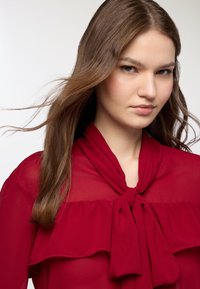 Blusa rossa trasparente con collo a fiocco e design a strati con volants. Il tessuto appare leggero, con una consistenza liscia e una caduta morbida.