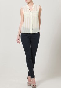 Blusa senza maniche a strisce giallo chiaro e bianco, leggera e trasparente, con colletto, abbinata a jeans skinny scuri e sandali con tacco alto grigio chiaro.