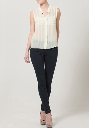 Blusa senza maniche a strisce giallo chiaro e bianco, leggera e trasparente, con colletto, abbinata a jeans skinny scuri e sandali con tacco alto grigio chiaro.