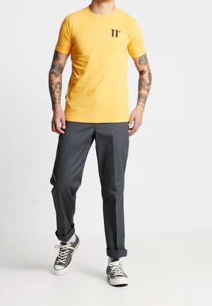 Gelbes T-Shirt mit kurzen Ärmeln und schwarzem "11°"-Logo, kombiniert mit grauer Hose und grünen Sneakern. Glatter Stoff, lässige Passform.