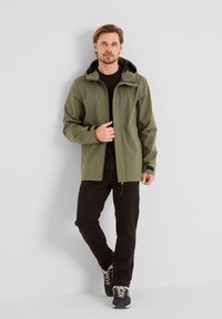 Olive green waterproof jacket with a hood, zip closure, elastic cuffs, and black accents; worn over a black shirt and paired with black pants.