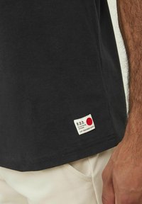 T-shirt di cotone nera con una piccola etichetta bianca che presenta un cerchio rosso e testo all'orlo, abbinata a pantaloni di colore chiaro.