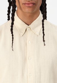 Flødefarvet skjorte med knapper og et tekstureret mønster af lodrette striber, med klassisk krave og hvide knapper, synlige på brystet.