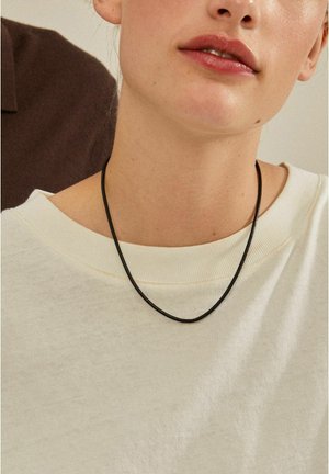 Black leather necklace worn over a cream-colored, textured sweater with a wide neckline, highlighting the contrast in color and materials.