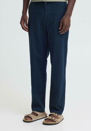 Mann trägt marineblaue Hose und beige Wildledersandalen mit zwei Schnallen, steht vor einem schlichten hellen Hintergrund.