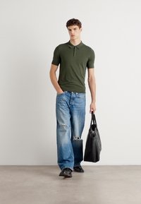 Olivengrønn kortermet polo skjorte kombinert med lyseblå slitte jeans og svarte sko. Modellen bærer en svart tote bag.