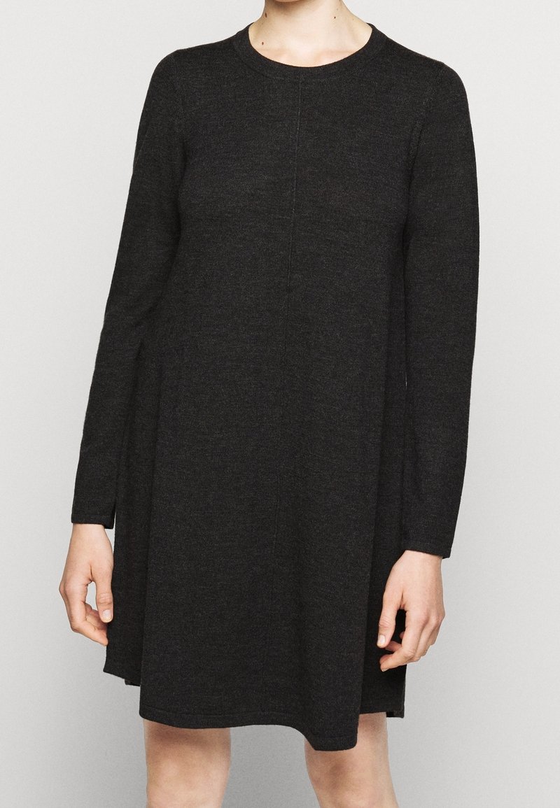 Femme portant une robe noire à manches longues et amples avec un col rond, debout devant un fond clair uni.