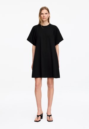 Femme portant une robe noire ample à manches courtes et des sandales à talons noires, debout devant un fond blanc uni.
