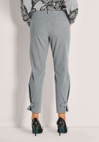 Vrouw in lichtgrijze op maat gemaakte broeken met achterzakken en gespdetails aan de pijpen, gecombineerd met zwarte hoge hakken.