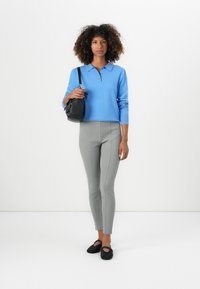 Lichtblauwe long-sleeve polo, grijze fitted leggings, zwarte crossbody tas, zwarte textuur flats, staand tegen een witte achtergrond.