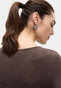 Brun struktur sweater med rund udskæring, parret med skinnende sølv kugleøreringe. Håret er stylet i en hestehale med et sort elastik.