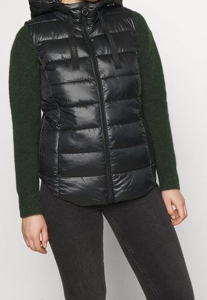 Zwarte doorgestikte mouwloze puffer-vest met ritssluiting en capuchon, gedragen over een donkergroene trui met lange mouwen door een persoon die staat.