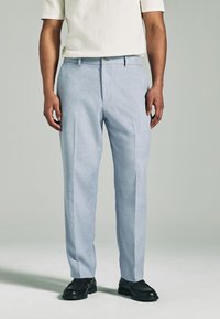 Himmelblaue maßgeschneiderte Hosen mit glatter Textur, ausgestattet mit Fältchen und klassischem Design. Kombiniert mit schwarzen Schuhen für einen Kontrast.