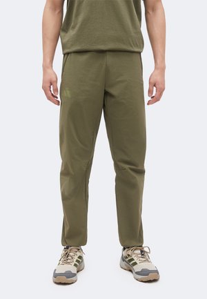 Homme portant un pantalon décontracté vert olive et une chemise vert olive avec des baskets beige et vertes, debout devant un fond uni clair.