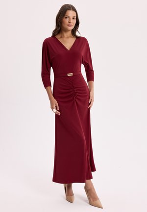 Kobieta stojąca w długiej bordowej sukni z rękawami do łokcia, marszczeniem na przodzie, paskiem w talii i beżowymi szpiczastymi szpilkami.