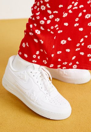 Hvide Nike sneakers med prægede blomsterdesigns, båret med hvide sokker og røde bukser med små hvide blomster på en gul gulv.