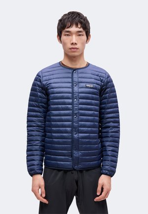 Giovane uomo che indossa una giacca trapuntata blu navy con bottoni e logo Patagonia sul petto sinistro, in piedi davanti a uno sfondo chiaro e semplice.
