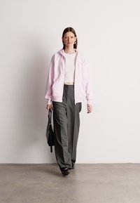 Svetlo ružová mikina na zips cez biely crop top, v kombinácii so šedými nohavicami s prúžkami a širokými nohavicami. Model drží čiernu tašku. Hladký, neformálny štýl.