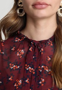 Blusa bordeaux con stampa floreale in bianco, rosso e blu navy. Presenta un colletto con lacci e dettagli arricciati all'escursione. Orecchini circolari dorati.