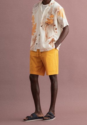 Hombre con camisa de manga corta beige con estampado de conchas marinas, pantalones cortos amarillos mostaza y sandalias negras, de pie frente a un fondo malva.