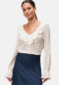 Kanten blouse in crème met een V-hals en ruches. Lange mouwen hebben gegolfde randen en zijn gecombineerd met een donkere spijkerrok.