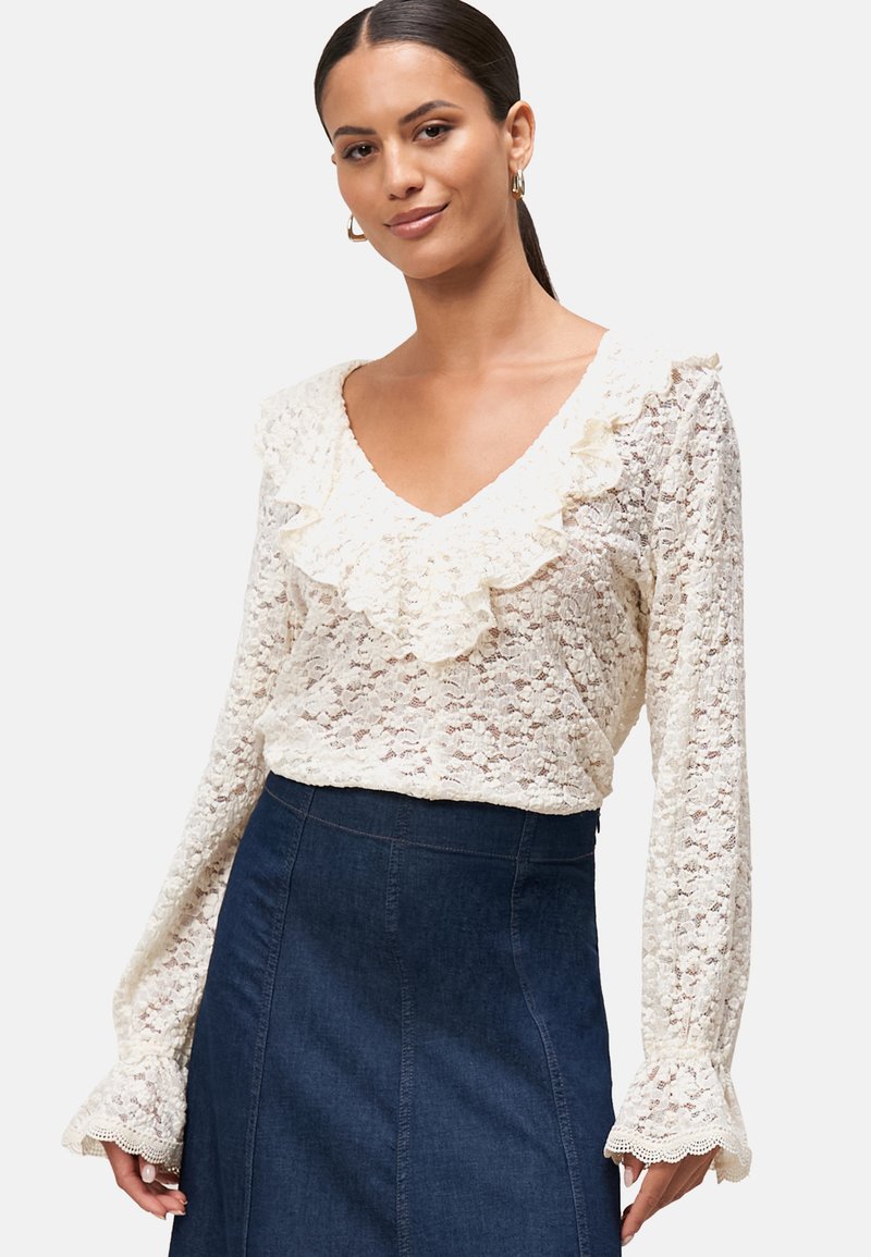 Kanten blouse in crème met een V-hals en ruches. Lange mouwen hebben gegolfde randen en zijn gecombineerd met een donkere spijkerrok.