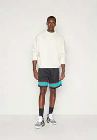 Balta sporta krekls, melni sporta šorti ar ziliem un sarkanajiem akcentiem, un pelēki sporta apavi. Stāv pret vienkāršu fonu, rokas atpūstās.