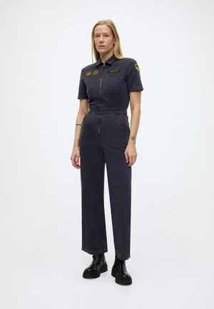 Femme debout en combinaison sombre à manches courtes, avec des écussons sur la poitrine et le bras, portée avec des bottes noires massives, sur un fond uni.