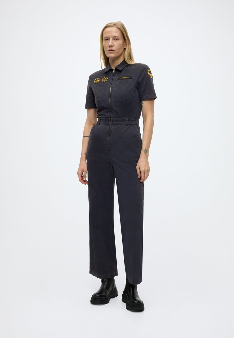 Femme debout en combinaison sombre à manches courtes, avec des écussons sur la poitrine et le bras, portée avec des bottes noires massives, sur un fond uni.