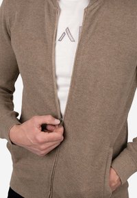 Cárdigan beige con cremallera, tejido texturizado, puños acanalados y bolsillos. Llevado sobre una camisa blanca con un patrón geométrico azul.