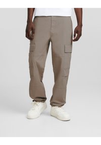 Beiget väriset cargo-housut, joissa on useita taskuja, rento istuvuus ja joustavat lahkeensuut, yhdistettynä valkoisiin tennareihin. Sileä kangaspinta.