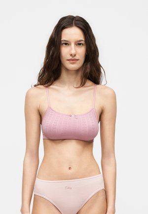 Femeie cu păr lung și castaniu care poartă un bralette roz din dantelă și lenjerie roz deschis cu textul „Friday” pe un fundal simplu.