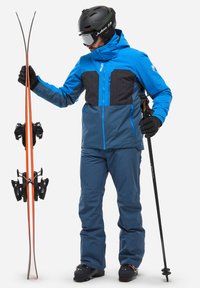 Skiausrüstung mit einer blauen und grauen Jacke, die ein strukturiertes schwarzes Panel aufweist. Blaue Hose, schwarze Handschuhe, Helm und orangefarbene Skier mit Bindungen.