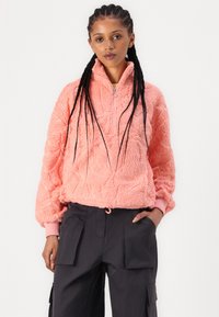 Vaaleanpunainen fleece-pullove, jossa on korkea kaulus, puolivetoketju, teksturoitu kuvio ja resorit, yhdistettynä tummiin cargo-housuihin, joissa on sivutaskut.