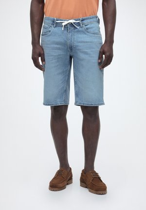 Mężczyzna w jasnoniebieskich dżinsowych szortach z białym sznurkiem, brązowych skórzanych butach i pomarańczowej koszuli, stojący na jednolitym tle.