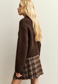 Donna bionda con capelli lunghi che indossa un maglione di lana marrone e una gonna plissettata a quadri marrone, girata di lato su uno sfondo neutro.