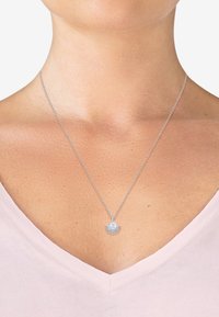 Collana d'argento con una delicata catena e un pendente a raggi di sole con un centro iridescente, indossata su una blusa a scollo a V.