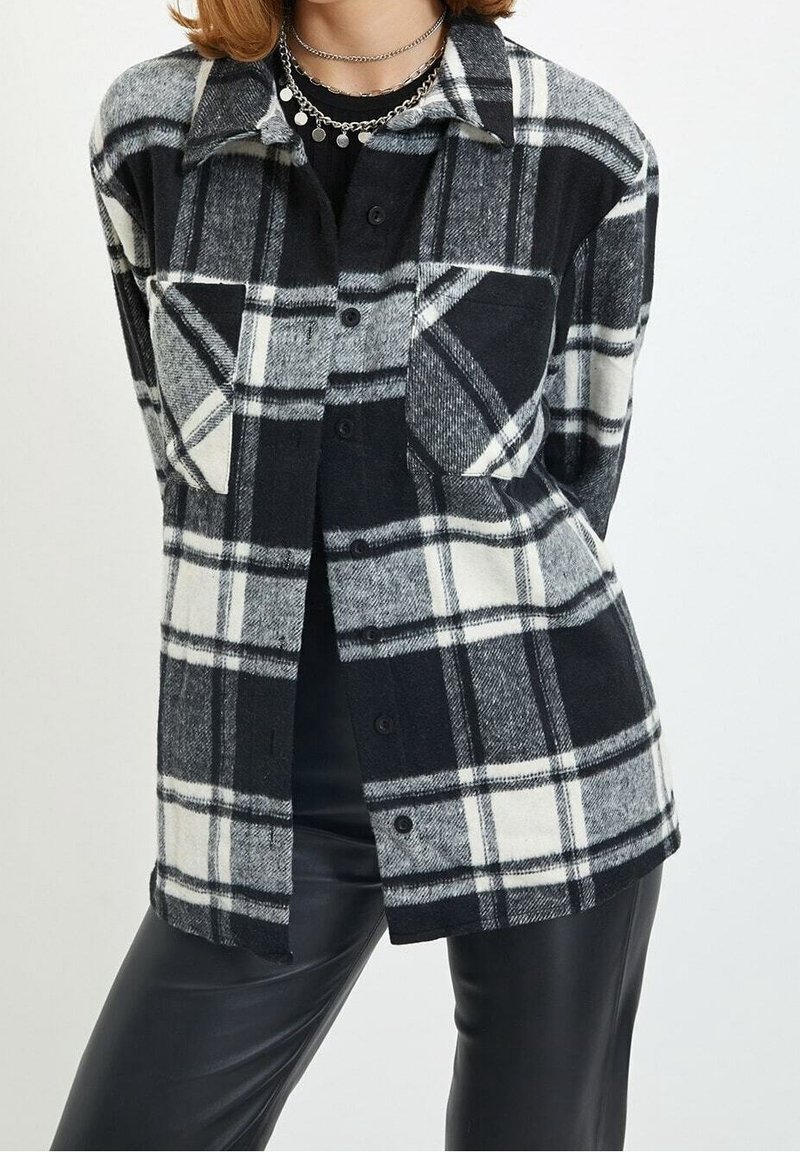 Camicia di flanella a quadri in bianco e nero con chiusura a bottoni e due tasche sul petto. Ha un colletto classico e un design con vestibilità ampia.
