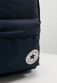 Tmavě modrý látkový batoh má kulatou bílou nášivku s červeným písmem a modrou hvězdou. Jednoduchý design s vyztuženým švem a elegantním tvarem.