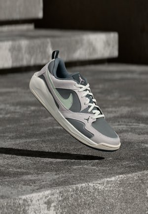 Pelēks un balts Nike Air Jordan sporta apavu pāris ar gaiši zaļu svītru, kas peld virs betona virsmas ar pakāpieniem fonā.